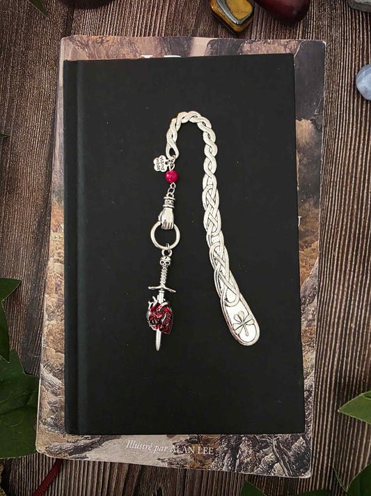 Marque-page "Le voeu de l'épée", un coeur humain transpercé par une épée, avec une breloque main au dessus qui tient l'épée ainsi qu'une pierre naturelle de corail rouge. Le signet est en métal et évoque un bijou de livre très beau pour les romans de fantasy, dark fantasy et romantasy