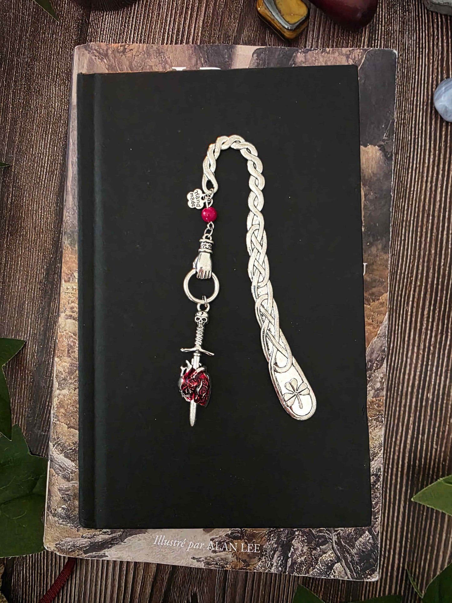 Marque-page "Le voeu de l'épée", un coeur humain transpercé par une épée, avec une breloque main au dessus qui tient l'épée ainsi qu'une pierre naturelle de corail rouge. Le signet est en métal et évoque un bijou de livre très beau pour les romans de fantasy, dark fantasy et romantasy