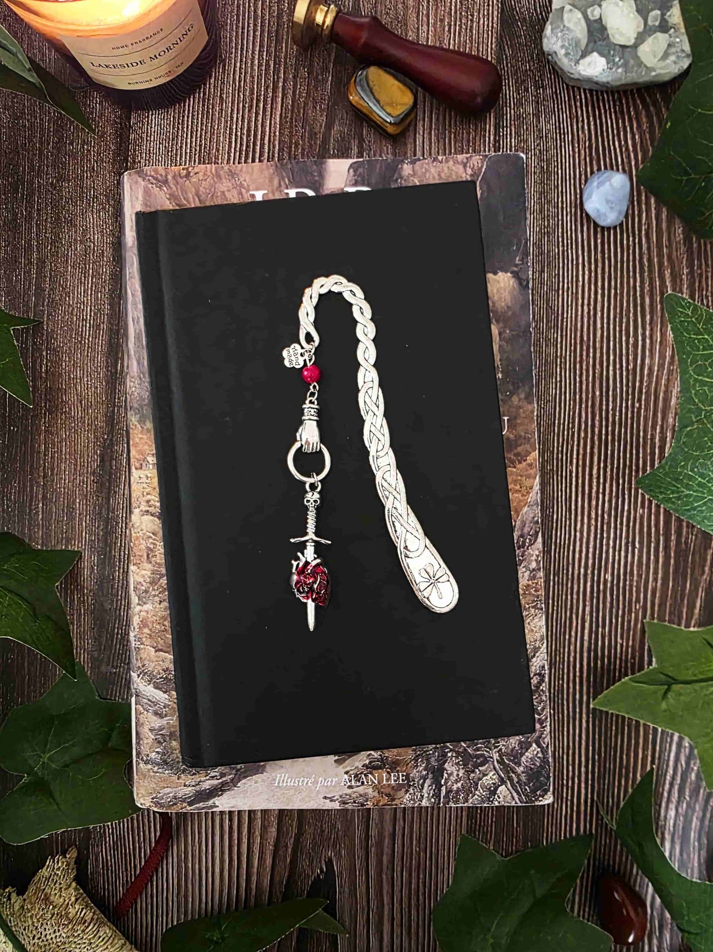Marque-page "Le voeu de l'épée", un coeur humain transpercé par une épée, avec une breloque main au dessus qui tient l'épée ainsi qu'une pierre naturelle de corail rouge. Le signet est en métal et évoque un bijou de livre très beau pour les romans de fantasy, dark fantasy et romantasy