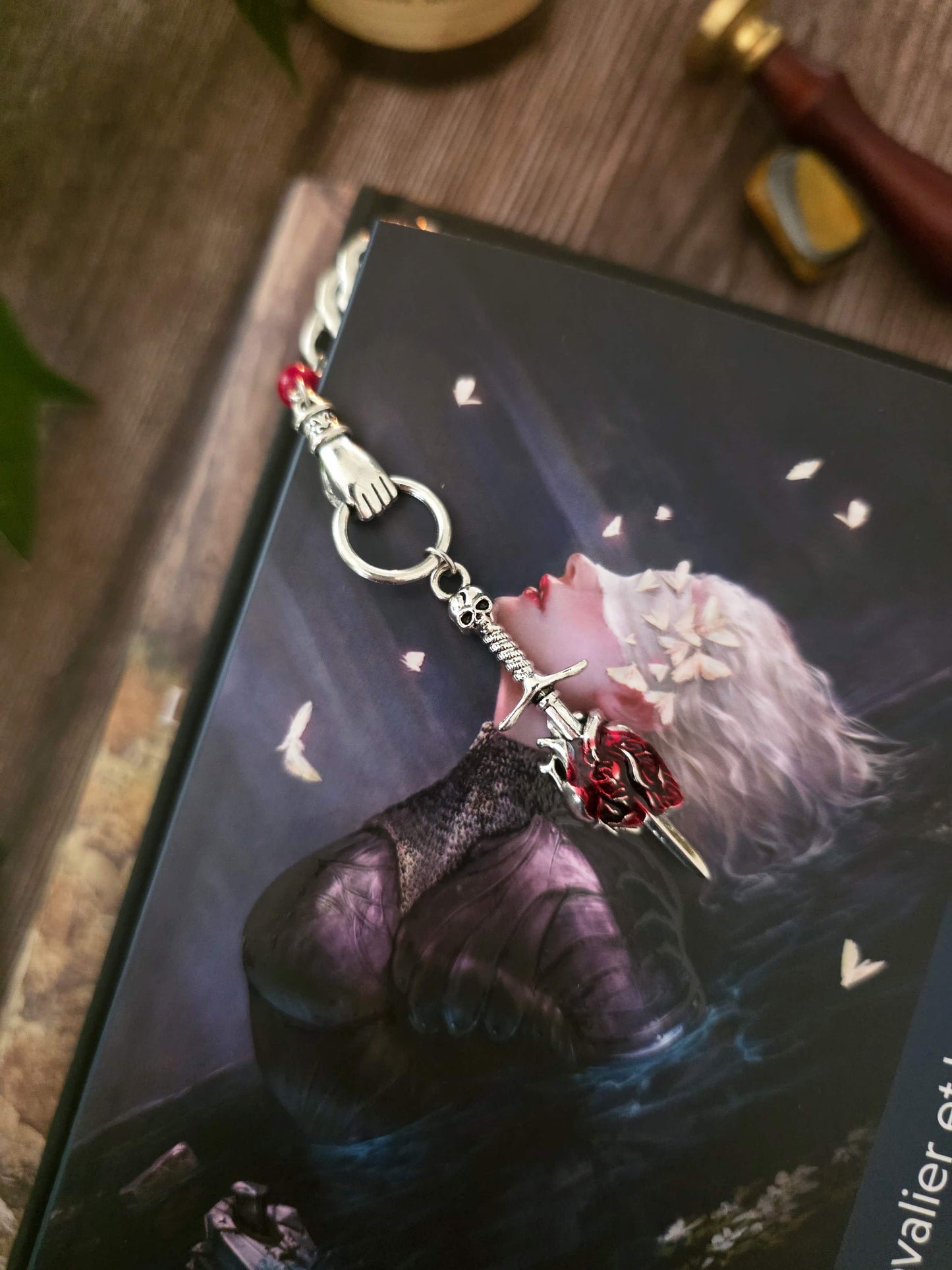 Marque-page "Le voeu de l'épée", un coeur humain transpercé par une épée, avec une breloque main au dessus qui tient l'épée ainsi qu'une pierre naturelle de corail rouge. Le signet est en métal et évoque un bijou de livre très beau pour les romans de fantasy, dark fantasy et romantasy