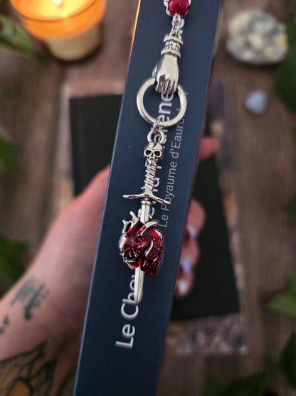Marque-page "Le voeu de l'épée", un coeur humain transpercé par une épée, avec une breloque main au dessus qui tient l'épée ainsi qu'une pierre naturelle de corail rouge. Le signet est en métal et évoque un bijou de livre très beau pour les romans de fantasy, dark fantasy et romantasy. dans un livre