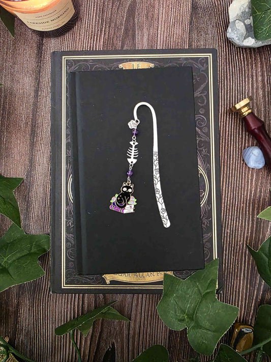 Marque-page en métal d'un chat noir assis sur une pile de livres violets évoquant des grimoire. Une arête de poisson est en breloque au centre et il y a deux pierres naturelles d'améthyste de chaque côté. Le bijou de livre est posé sur un livre noir 